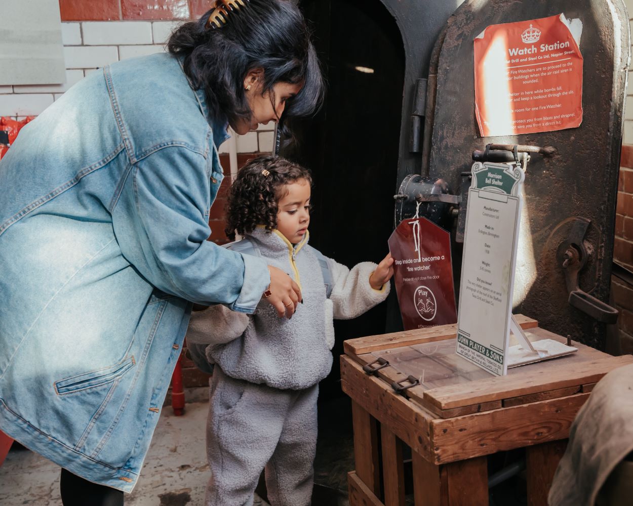 A young girl and a woman look at a Fire Watch station exhibit at the National Emergency Services Museum. The girl is touching a label hanging from an object.