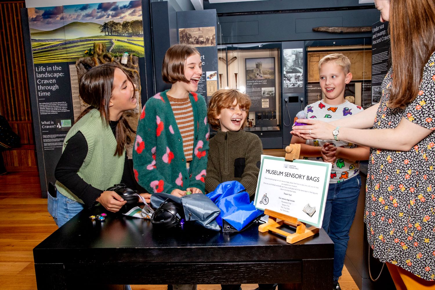 A group of four children gathered round a desk filled with objects and a sign reading museum sensory bags. A museum staff member is holding out a toy to them and they are looking at it and laughing.