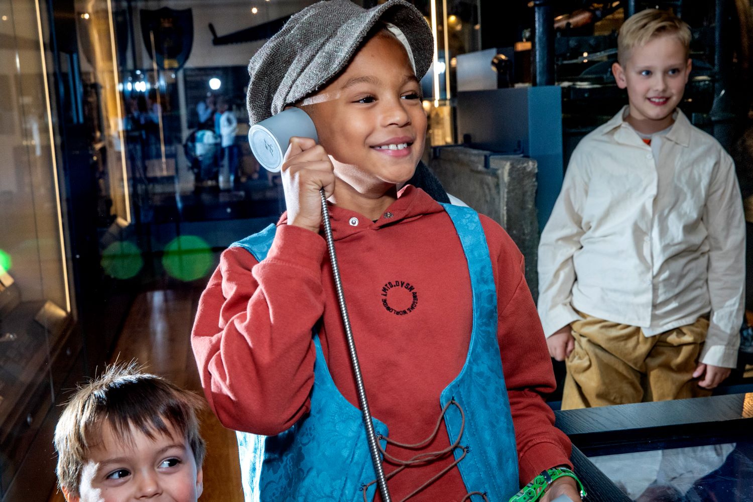 A young child wearing a flat cap and blue waistcoat on top of his clothes holds a speaker up to his ear and grins at Craven Museum.