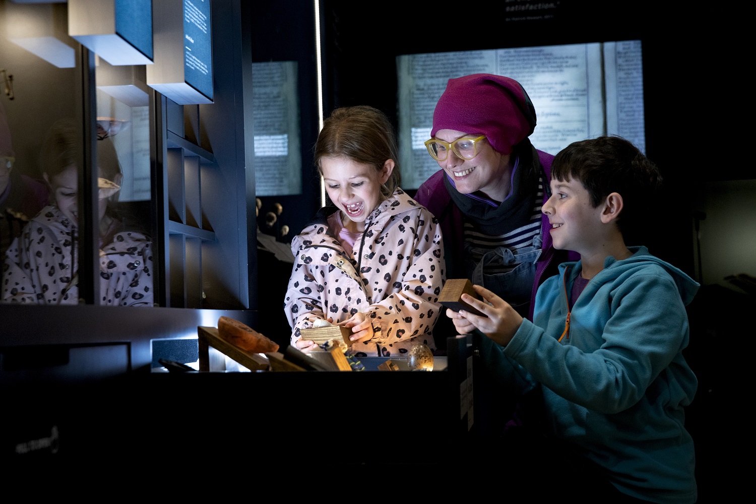 A woman and two children smiling in wonder in a gallery at Craven Museum. The two children look and at hold up objects from a pull out drawer in the gallery.