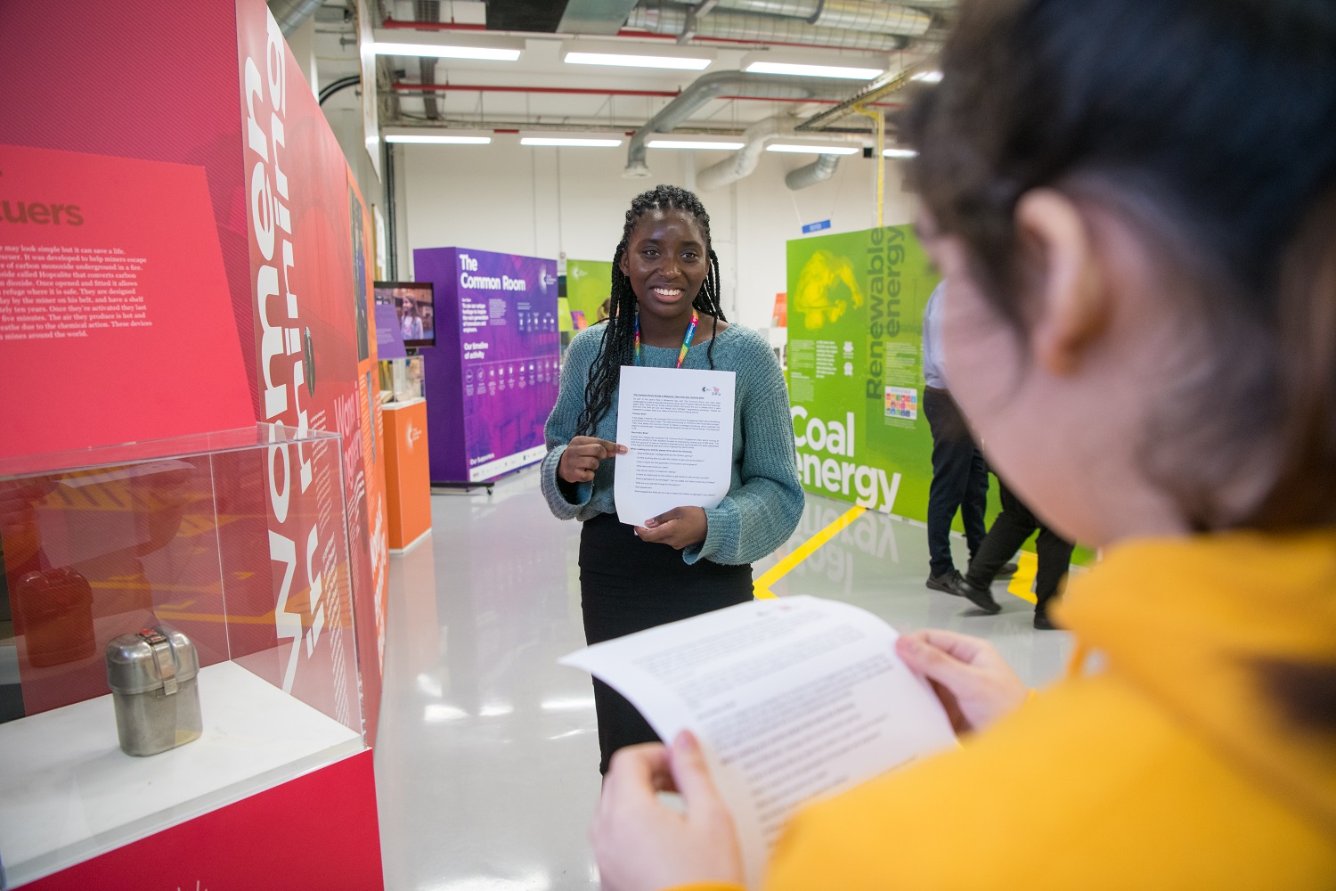 A young woman smiles and stands in front of another holding an A4 sheet of paper in an exhibition at the Common Room and Caterpillar Takeover Day.