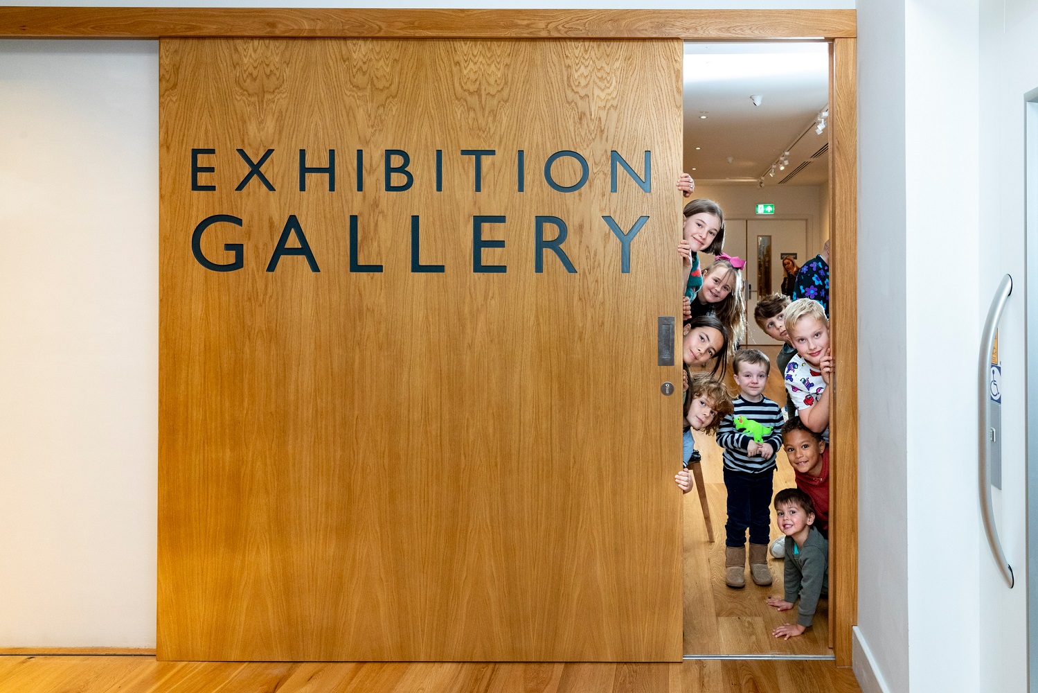 A group of children grin and peer round a gap in a wooden sliding door reading Exhibition Gallery at Craven Museum.