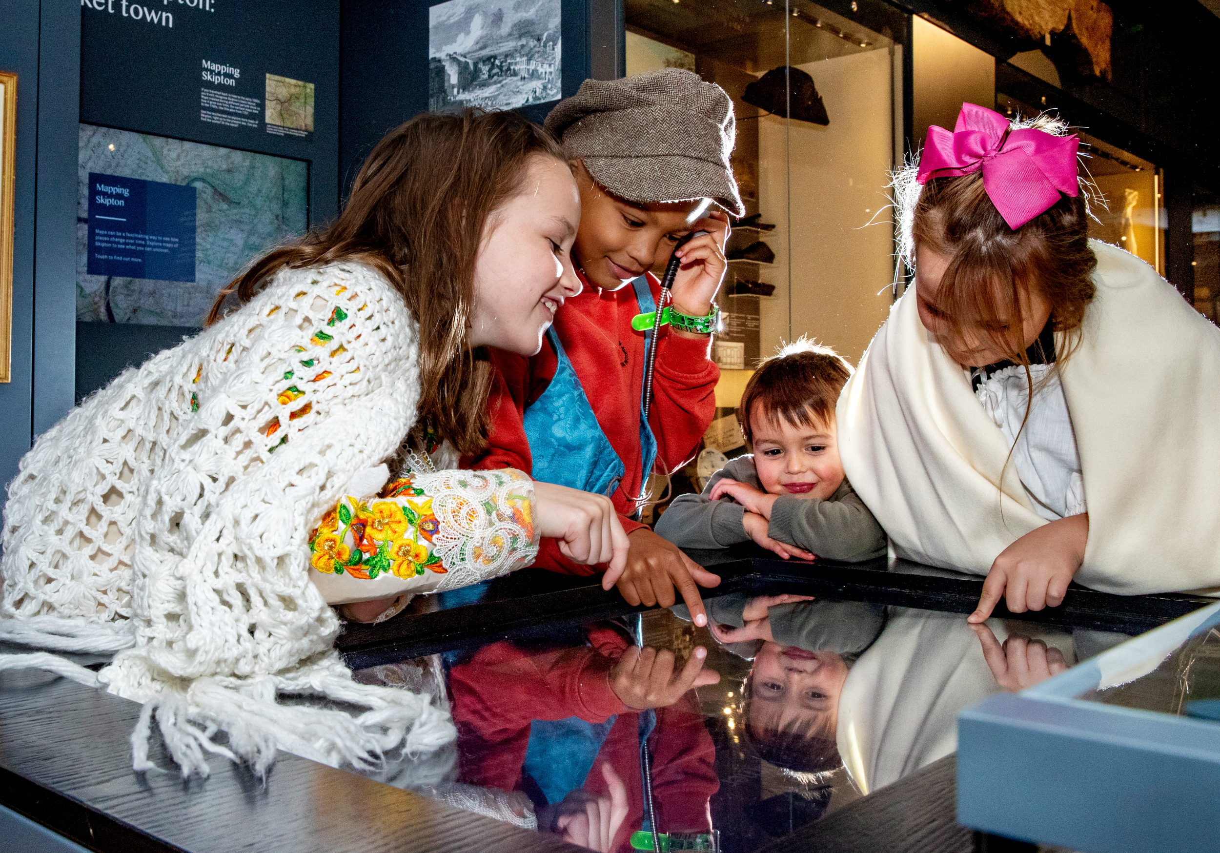 A group of four children dressed in different museum costumes lean over an interactive display screen at Craven Museum. One holds a speaker up to his ear.