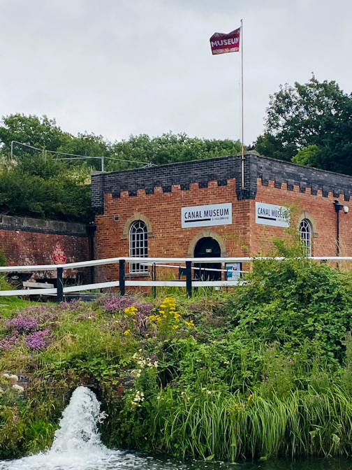 In the foreground, there are bushes and wild flowers with a small fountain in the middle. Behind the bushes is a small brick building with signs saying Canal Museum. There is a flag flying above the building which says museum