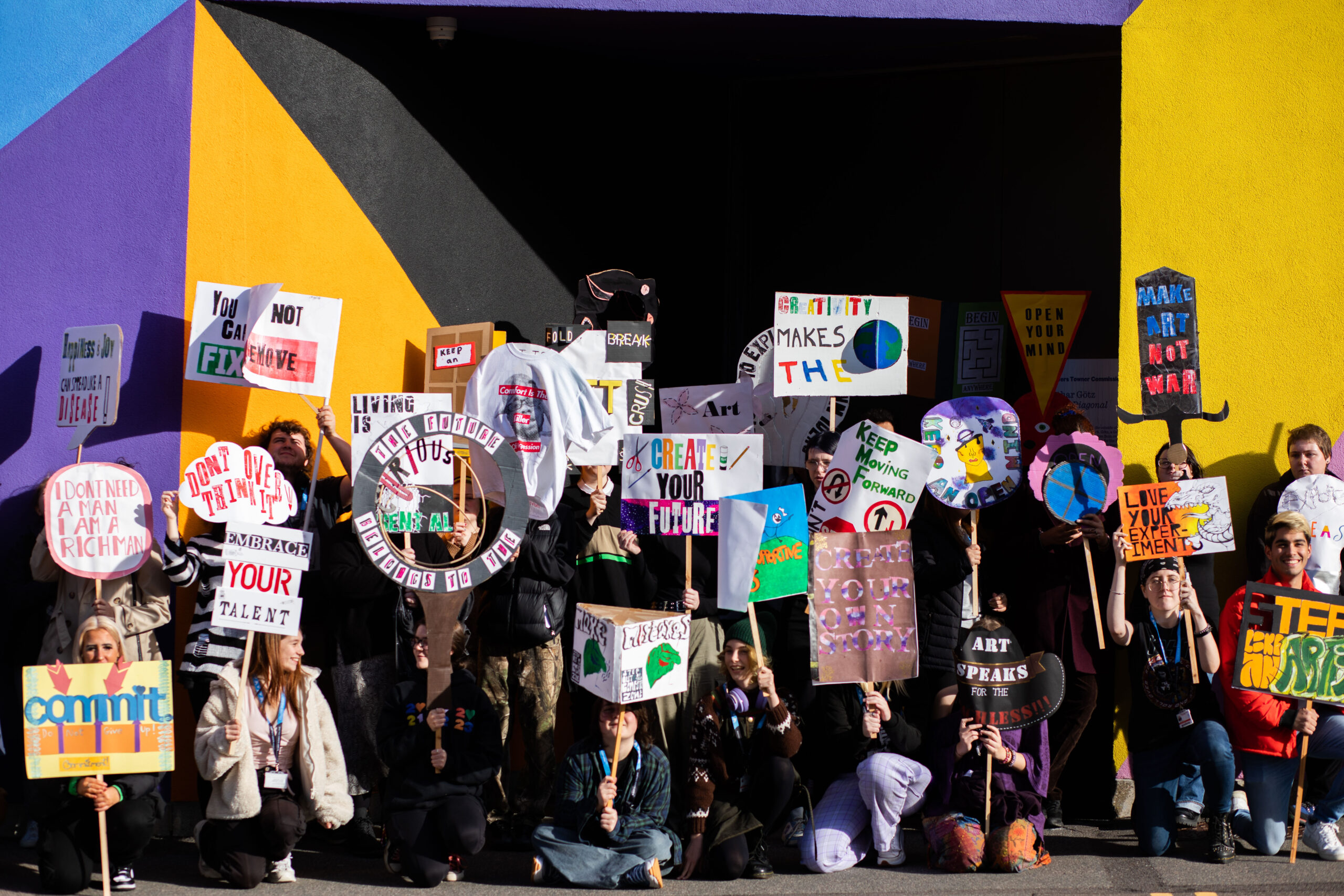 A group of young people stand outside a building on a sunny day holding protest placards, with a range of phrases including: Make Art Not War, Creativity Makes the Earth, Embrace Your Talent.