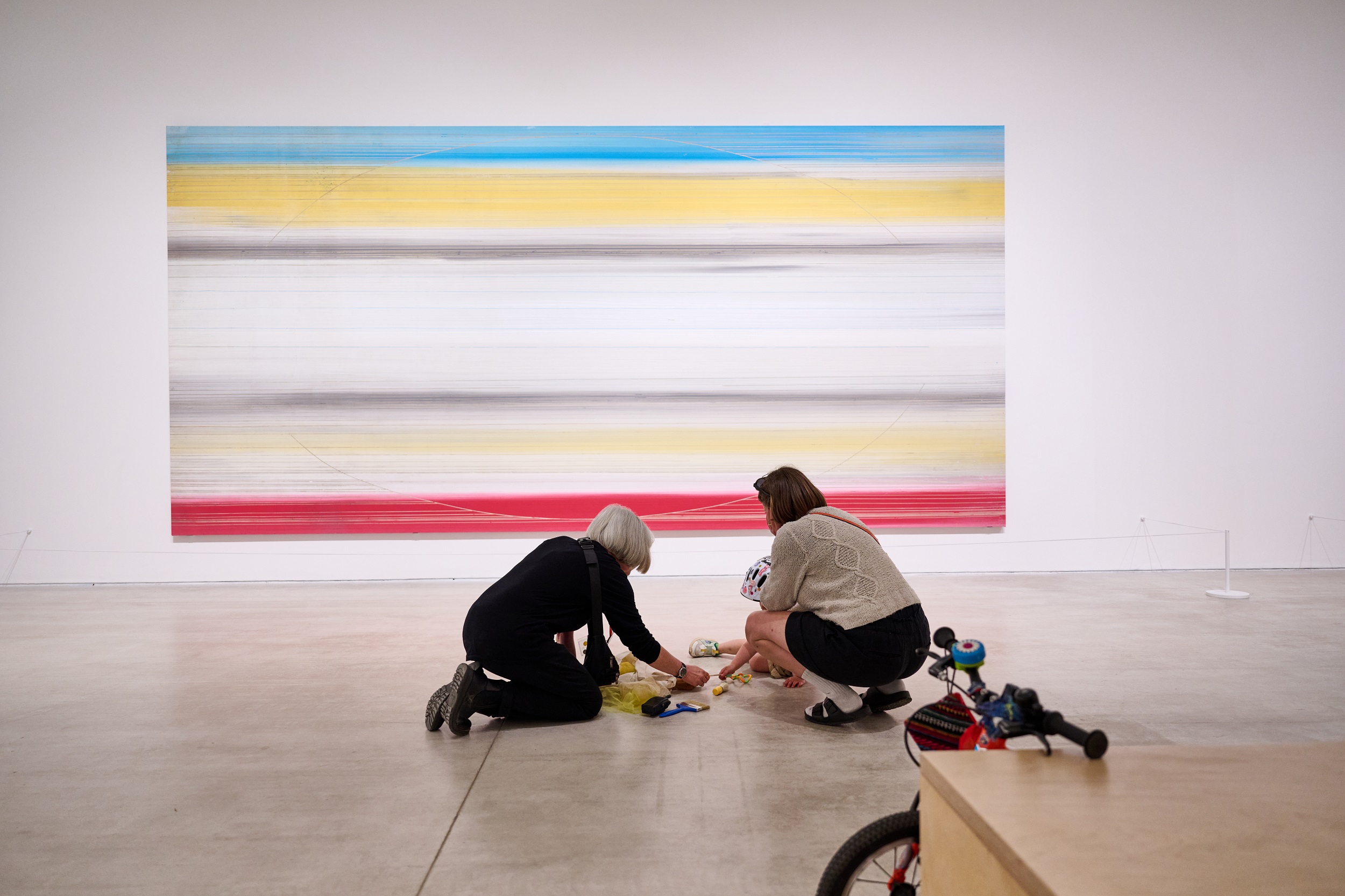 Two adults, one with grey hair, and a young child sit on the floor of a large gallery space in front of a colourful abstract painting. They are playing with the contents of a sensory bag. A bike is propped up against a bench in the foreground.